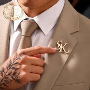 Könnte beinhalten: Goldfarbene Anstecknadel mit den Initialen "SK" an einem beigefarbenen Anzug befestigt. Die Person trägt ein weißes Hemd und eine beige Krawatte. Ein Tattoo eines Blattzweigs ist auf der Hand zu sehen.