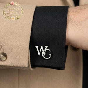 Op de afbeelding: Een zilverkleurige manchetknop met de in elkaar grijpende letters WG, bevestigd aan de zwarte stoffen manchet van een overhemdmouw, die gedeeltelijk wordt bedekt door een beige blazer-mouw met een zichtbare knoop.