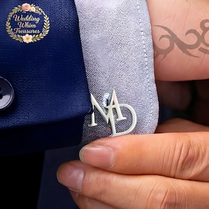 Op de afbeelding: Zilveren manchetknopen met de initialen "ND" worden getoond op een lichtblauwe overhemdmouw. De manchetknopen bevinden zich naast een donkerblauwe colbertjas. De afbeelding toont ook een tatoeage op de arm.
