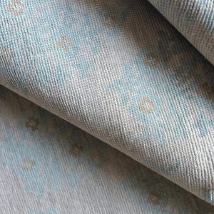 Pode incluir: Close-up de um tapete enrolado em tons de azul claro e branco sujo. O tapete apresenta um padrão repetitivo de pequenos motivos florais estilizados em cor dourada suave. A textura parece macia e tecida.