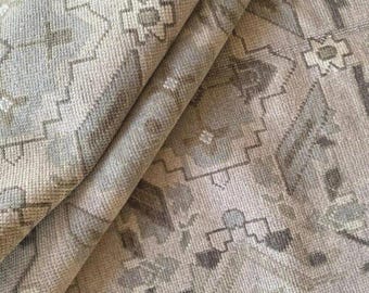 May include: A close-up shot of a folded rug with a muted colour scheme. The rug displays a repeating geometric pattern in beige, grey, and muted green. The texture is visible, with a slightly worn look. The design incorporates floral and angular shapes.