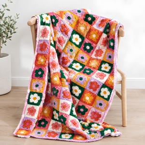 Op de afbeelding: Een handgemaakte gehaakte deken met een herhalend patroon van vierkante motieven, elk met een veelkleurig bloemendesign. De deken gebruikt een levendig palet van oranje, geel, roze, wit, paars en donkergroen garen door het hele ontwerp.