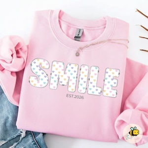 Puede incluir: Sudadera rosa claro con la palabra "SMILE" en una fuente juguetona. Las letras están rellenas de corazones pastel y delineadas con un diseño de puntada punteada. El texto "EST.2026" está debajo.