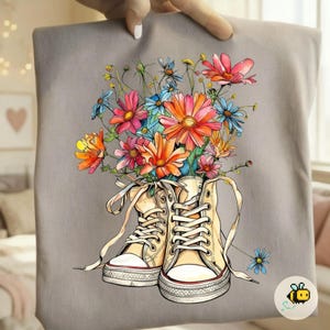 May include: A gray tote bag featuring a colorful floral design. The artwork depicts a pair of cream-colored high-top sneakers filled with a vibrant bouquet of pink, orange, and blue flowers. The bag is held by a person.