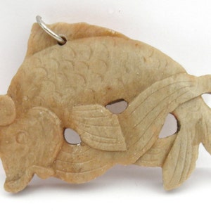 May include: A carved jade pendant in the shape of a fish. The pendant is a light brown color and has a hole at the top for a cord or chain.