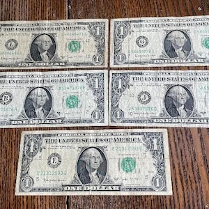 May include: Five United States one-dollar bills are displayed on a wooden surface. Each bill features a portrait of George Washington and the text "The United States of America". The bills are in various conditions.