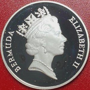 Może przedstawiać: Srebrna moneta z profilem królowej Elżbiety II, z napisem „ELIZABETH II” po prawej stronie i „BERMUDA” po lewej. Moneta ma czarne tło i srebrną obwódkę, na czerwonym tle.