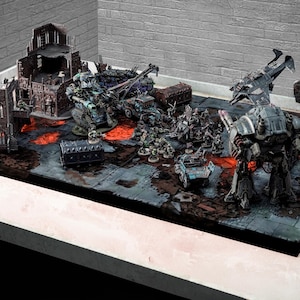 Puede incluir: Una detallada escena de juego de guerra de mesa con figuras en miniatura, vehículos y estructuras. El terreno incluye una superficie oscura y texturizada con elementos rojos similares a la lava. Varias figuras y vehículos están dispuestos en el tablero.