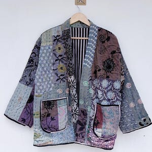 Peut inclure: Une veste en patchwork présentant une variété de motifs floraux et géométriques dans des tons de bleu, violet et gris. Le vêtement possède une ouverture frontale, des manches larges, deux grandes poches avant à bordure noire et une doublure rayée.