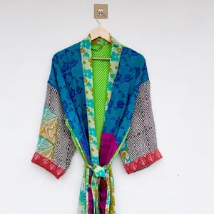 Pode incluir: Um roupão de manga comprida com um design de patchwork apresentando vários padrões florais e geométricos em tons de azul, verde, rosa e branco. A peça inclui um cinto de tecido a condizer na cintura e um forro interior às riscas verde brilhante.