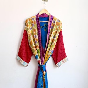 Può includere: Una vestaglia a maniche lunghe caratterizzata da un design patchwork con vari motivi e colori, tra cui rosso, giallo, blu e viola. Il capo presenta una chiusura con cintura in vita, un ampio colletto e una vivace struttura in tessuto multicolore.