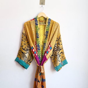Puede incluir: Una bata tipo kimono de manga larga con un diseño de retazos en tonos amarillo mostaza, naranja y verde azulado con motivos florales y geométricos. La prenda incluye un cinturón para anudar, mangas anchas y una solapa frontal en contraste.