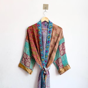 May include: A long-sleeved kimono robe featuring a patchwork design with various floral and geometric patterns in shades of pink, teal, blue, and tan. The garment includes a matching tie belt and a wide collar with a blue and white circular motif.