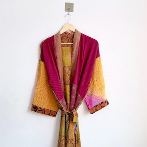 Puede incluir: Una bata de manga larga con un diseño de patchwork que presenta varios patrones de tela en tonos magenta, amarillo mostaza y marrón. La prenda incluye un cuello ancho, un cinturón para anudar a la cintura y un dobladillo bajo las rodillas.