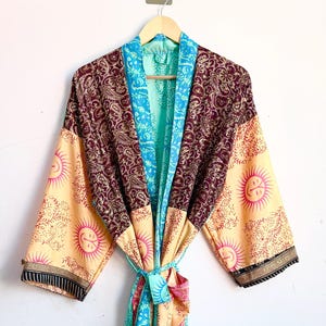 Op de afbeelding: Een kamerjas met lange mouwen en een patchwork-ontwerp, bestaande uit crèmekleurige panelen met roze zonmotieven, donkerbordeauxrode delen met complexe paisley-patronen en helder turquoise randen met gedetailleerde bloemenprints.