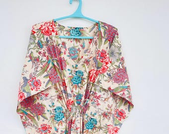 May include: A cream-coloured kaftan with a vibrant floral pattern in red, pink, and teal. The lightweight garment features a loose, flowing design with wide sleeves and a drawstring waist. The kaftan is hanging on a blue hanger against a white wall.