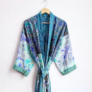 May include: A long-sleeved kimono robe featuring a vibrant patchwork of blue, teal, and turquoise paisley patterns. The garment includes a matching fabric tie belt at the waist, wide sleeves, and a shawl collar, all displayed on a wooden hanger.