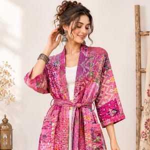 May include: A long-sleeved, knee-length patchwork duster coat in vibrant shades of pink, magenta, and purple. The garment features a tie-waist belt, two front pockets, and a variety of intricate, multi-colored floral and geometric patterns.