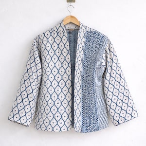 Könnte beinhalten: Gesteppte Jacke mit weißem und blauem Rautenmuster. Die Jacke hat lange Ärmel und eine kontrastierende blau gemusterte Seite. Sie hängt an einem Holzbügel vor einer weißen Wand.