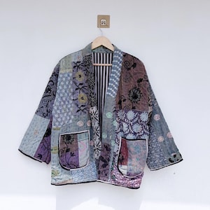 Puede incluir: Una chaqueta de patchwork con una variedad de patrones florales y geométricos en tonos azules, morados y grises. La prenda tiene un frente abierto, mangas anchas, dos bolsillos delanteros grandes con ribete negro y un forro interior a rayas.