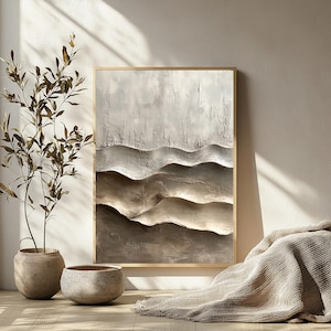 May include: Textured abstract wall art in a gold frame. The artwork features a layered design with shades of gray and brown, creating a sense of depth and dimension. The piece is displayed against a neutral wall, with a plant and decorative pots in the foreground.