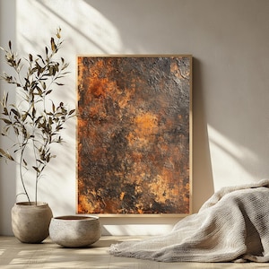 以下が含まれることがあります： 木製フレームに入った抽象芸術作品。このアートワークは、オレンジ、茶色、黒の色合いが混ざったテクスチャのある表面が特徴です。絵画は白い壁に立てかけられており、手前には植物と装飾的な鉢があります。