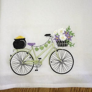 May include: A white kitchen towel with an embroidered design of a green bicycle with a basket of flowers and a pot of gold on the back rack.