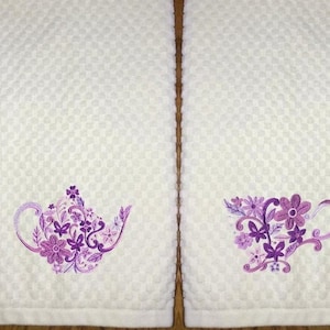 May include: Two white, textured hand towels with embroidered floral designs. One towel features a teapot, the other a teacup, both in shades of purple. The towels have a waffle weave pattern.