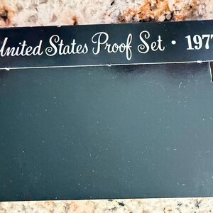 May include: A black rectangular cardboard box for a United States Proof Set from 1977. The top flap features white cursive and serif text that reads United States Proof Set • 1977. The box has a smooth finish and a tabbed closure design.