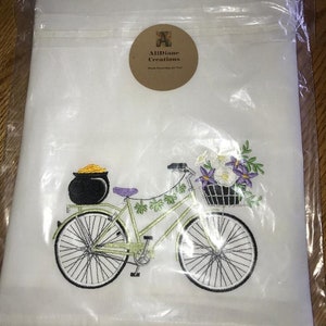 May include: White kitchen towel with an embroidered design of a green bicycle with a pot of gold on the back rack and a basket of flowers on the front. The bicycle has purple accents and is surrounded by shamrocks. The towel has a brown label with the text "Allidane Creations" and "Made Especially for You".