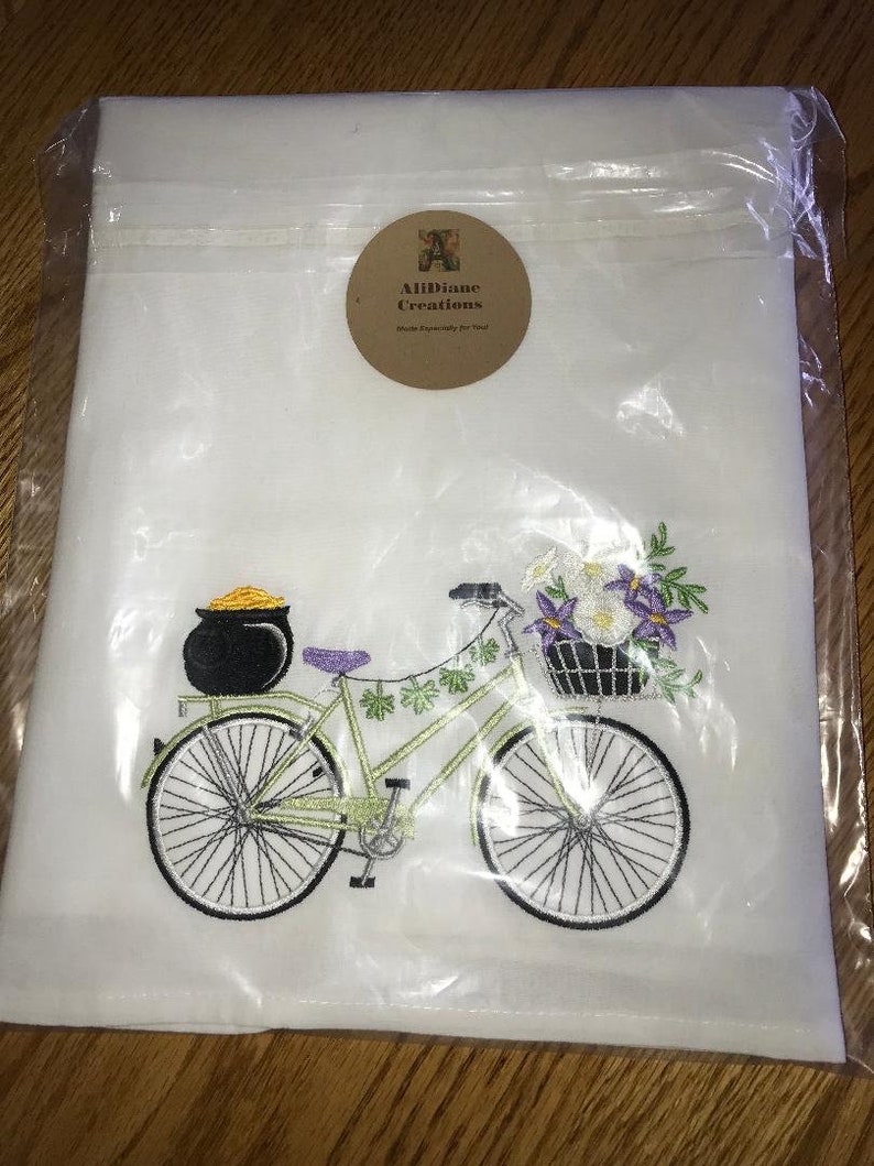 May include: White kitchen towel with an embroidered design of a green bicycle with a pot of gold on the back rack and a basket of flowers on the front. The bicycle has purple accents and is surrounded by shamrocks. The towel has a brown label with the text "Allidane Creations" and "Made Especially for You".