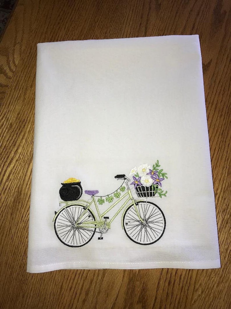 May include: A white kitchen towel with an embroidered design of a green bicycle with a basket of flowers and a pot of gold on the back rack.