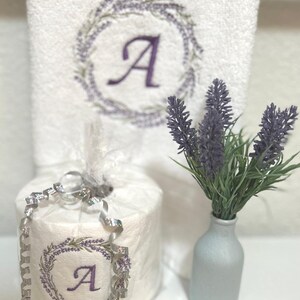May include: A white hand towel with a lavender wreath embroidered with the letter "A" in the center. A white toilet paper roll with a lavender wreath embroidered with the letter "A" in the center is wrapped in clear plastic with silver ribbon. A small vase with a bouquet of lavender flowers.