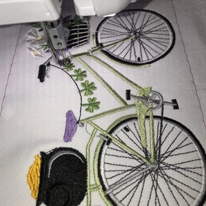 May include: A close-up of a green and black bicycle with a pot of gold at the front. The bicycle has a basket of flowers on the handlebars and is being embroidered on a white fabric.