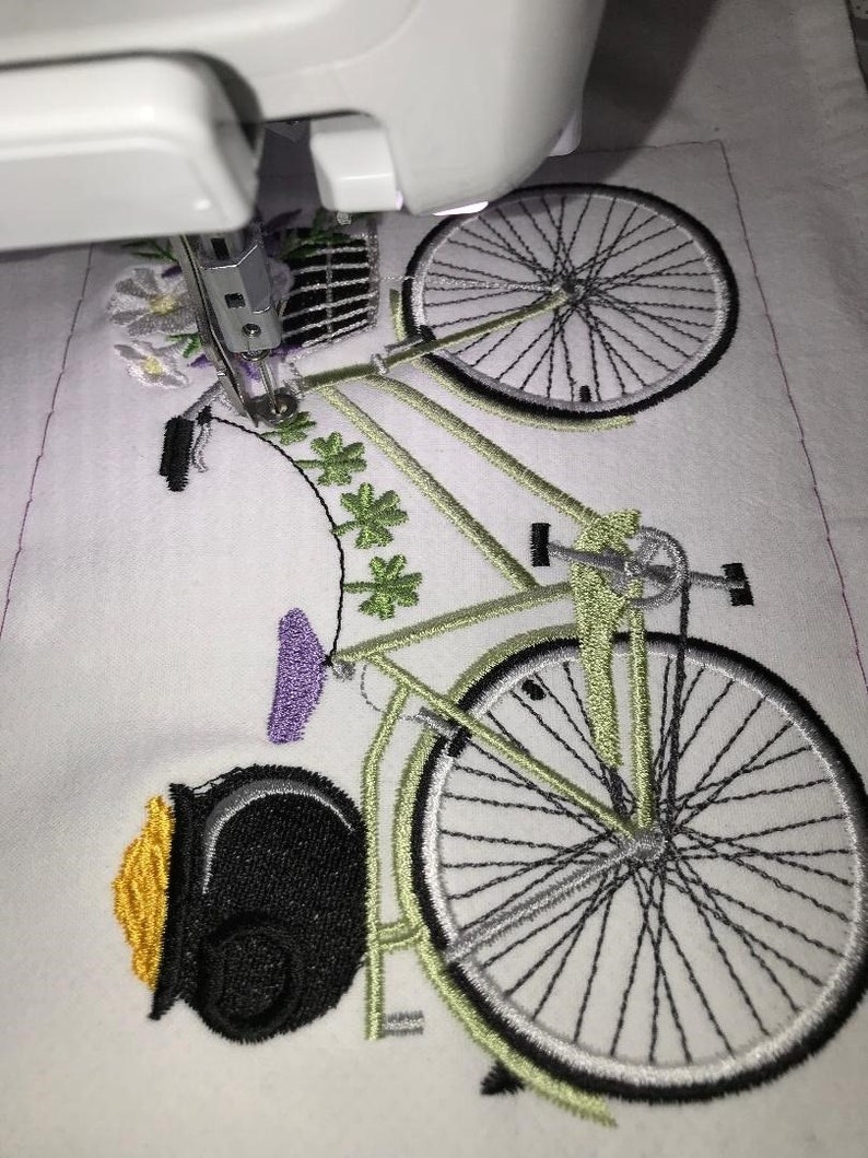 May include: A close-up of a green and black bicycle with a pot of gold at the front. The bicycle has a basket of flowers on the handlebars and is being embroidered on a white fabric.
