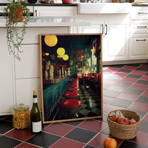 May include: A framed diner print leans against a white kitchen cabinet. The image features red diner stools, a teal counter, and yellow globe lights. A basket of apples, a wine bottle, and a jar of pasta are in the foreground.