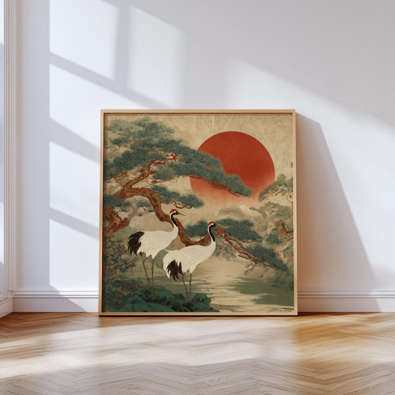 May include: Framed art print featuring a traditional Japanese landscape. Two white cranes with black and red markings stand near a body of water, beneath a large red sun and evergreen trees. The artwork is in a wooden frame, displayed against a neutral wall.