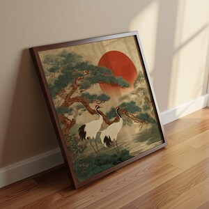May include: Framed artwork featuring two white cranes with black wingtips and red head markings, standing beneath a tree with a large red sun in the background. The artwork is in a dark brown frame, leaning against a wall.