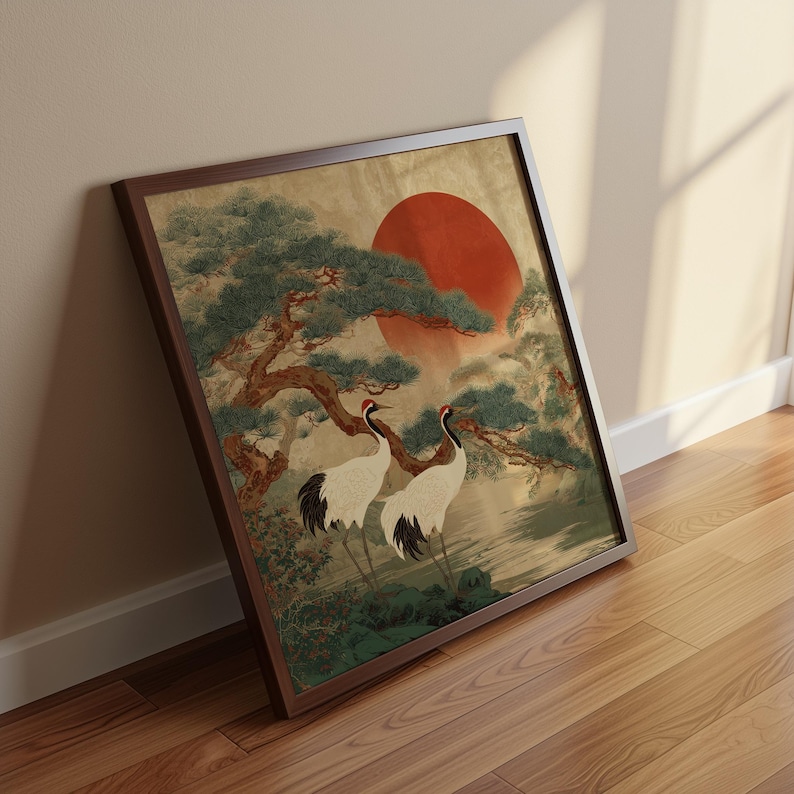 May include: Framed artwork featuring two white cranes with black wingtips and red head markings, standing beneath a tree with a large red sun in the background. The artwork is in a dark brown frame, leaning against a wall.