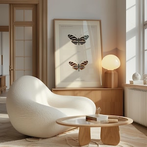 Puede incluir: Un estampado enmarcado con dos ilustraciones detalladas de mariposas en tonos negros y marrones. El estampado está colgado sobre un mueble de madera, junto a una silla blanca y redondeada y una mesa de madera con tapa de cristal. Una lámpara con una pantalla redonda y blanca está sobre el mueble.