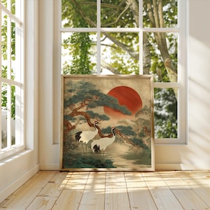 May include: Framed artwork featuring two cranes standing near water, with a large red sun in the background. The artwork has a traditional Japanese aesthetic, with a tree and foliage in shades of green and brown. The frame is a light brown color.