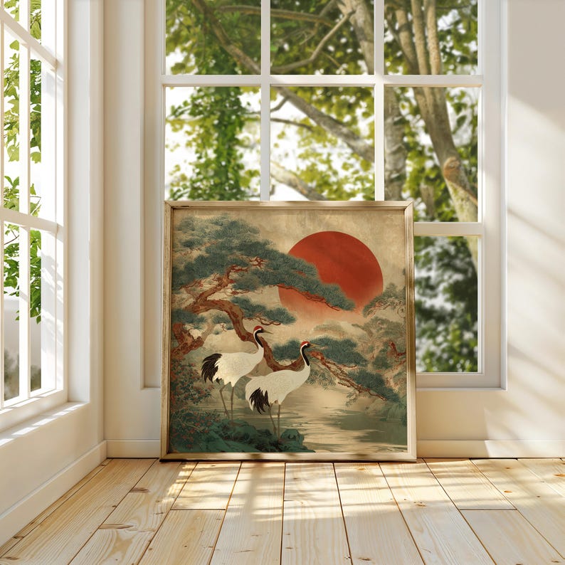May include: Framed artwork featuring two cranes standing near water, with a large red sun in the background. The artwork has a traditional Japanese aesthetic, with a tree and foliage in shades of green and brown. The frame is a light brown color.