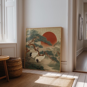 May include: Framed art print featuring a traditional Japanese landscape. The artwork depicts two cranes, a large red sun, and a pine tree. The color palette includes shades of green, red, and beige. The print is displayed in a room with white walls and a wooden floor.