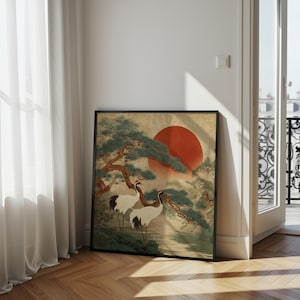 May include: Framed art print featuring a traditional Japanese landscape. The artwork depicts two white cranes with red crowns, a large red sun, and green trees. The print is in a black frame and leans against a wall.