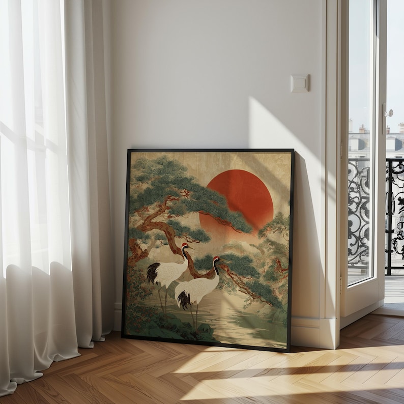 May include: Framed art print featuring a traditional Japanese landscape. The artwork depicts two white cranes with red crowns, a large red sun, and green trees. The print is in a black frame and leans against a wall.