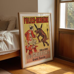 May include: Framed vintage poster for Folies-Bergère, featuring a clown and a donkey on a tightrope. The poster has a yellow background with red and black text. The clown is wearing a red and white costume. The donkey is brown.