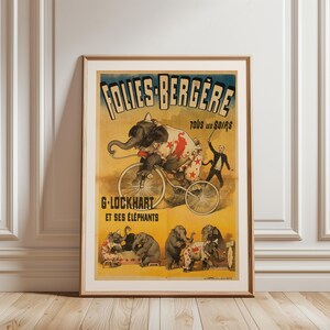 May include: Framed vintage circus poster featuring an elephant riding a bicycle, with text "FOLIES-BERGERE" and "G-LOCKHART ET SES ÉLÉPHANTS." The poster has a yellow background with illustrations of elephants in various circus acts. The frame is a light brown color.