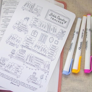 May include: A Bible reading tracker with a bookshelf design, featuring book titles and a decorative frame. Next to it are four Sharpie S-Note highlighters with blue, yellow, pink, and green accents. The tracker is open on a pink Bible.
