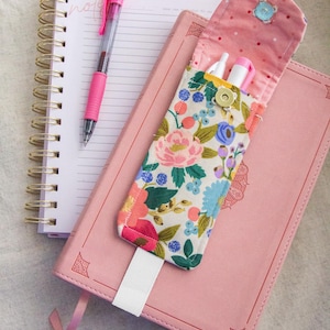 Könnte beinhalten: Ein rosa Stift liegt auf einem Spiralnotizbuch neben einem rosa Notizbuch mit einem Schleifenlesezeichen. Ein offenes Federmäppchen mit Blumenmuster und weißem Riemen zeigt Stifte und einen Textmarker.