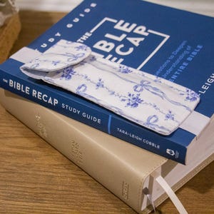 May include: A stack of books with a blue and white floral patterned case on top. The top book is titled "THE BIBLE RECAP STUDY GUIDE" in white text on a blue background. The bottom book is beige with a white ribbon bookmark.